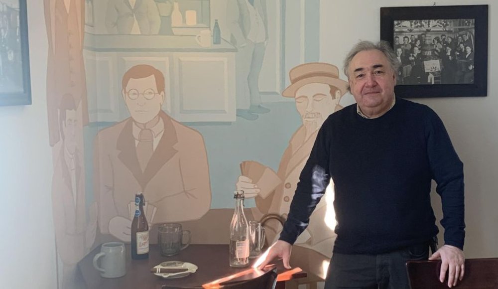 Ein Mann mit grauem Haar steht in einem Museumsraum neben einem Tisch und blickt in die Kamera. Hinter ihm ist eine großflächige Wandillustration zu sehen, die mehrere Personen in historischer Kleidung in einer Gaststube zeigt. Auf dem gemalten Tisch stehen Flaschen, Krüge und Geschirr. An der Wand hängt außerdem ein historisches Schwarz-Weiß-Foto mit einer Personengruppe.