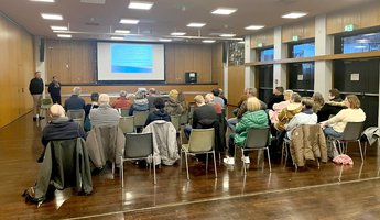 Personen sitzen vor auf Stühlen vor Leinwand mit Präsentation