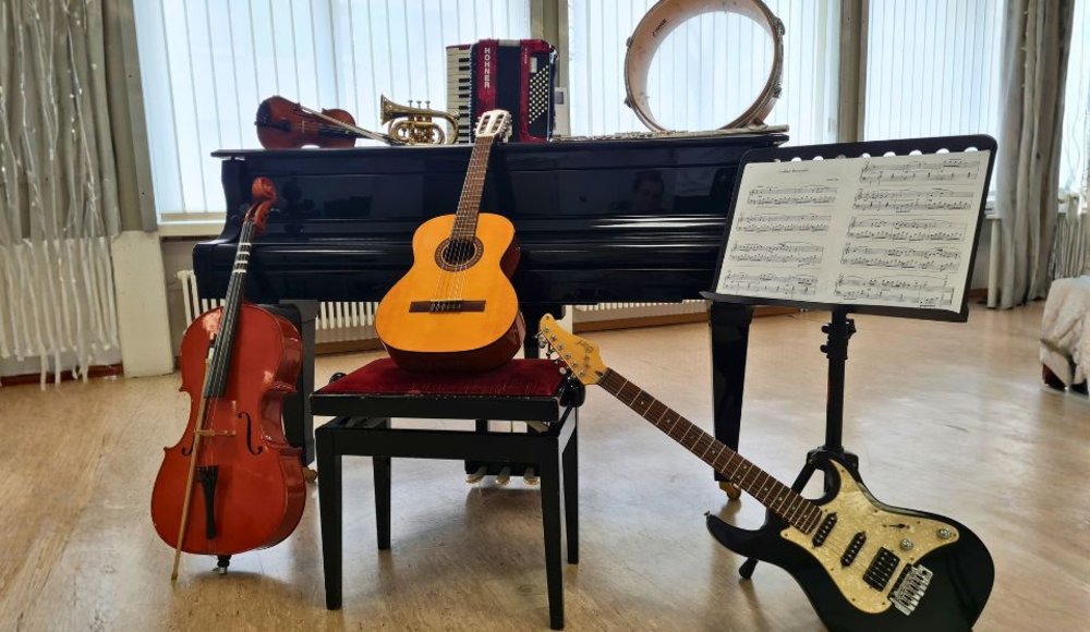 In einem hellen Musikraum steht ein schwarzer Flügel, vor dem verschiedene Musikinstrumente arrangiert sind: ein Cello, eine klassische Gitarre auf einem Klavierhocker, eine schwarze E-Gitarre, eine Geige, eine Trompete, ein Akkordeon und ein Tamburin. Rechts daneben befindet sich ein Notenständer mit aufgeschlagenen Noten. Das Foto vermittelt die Vielfalt des Unterrichtsangebots der Musikschule.