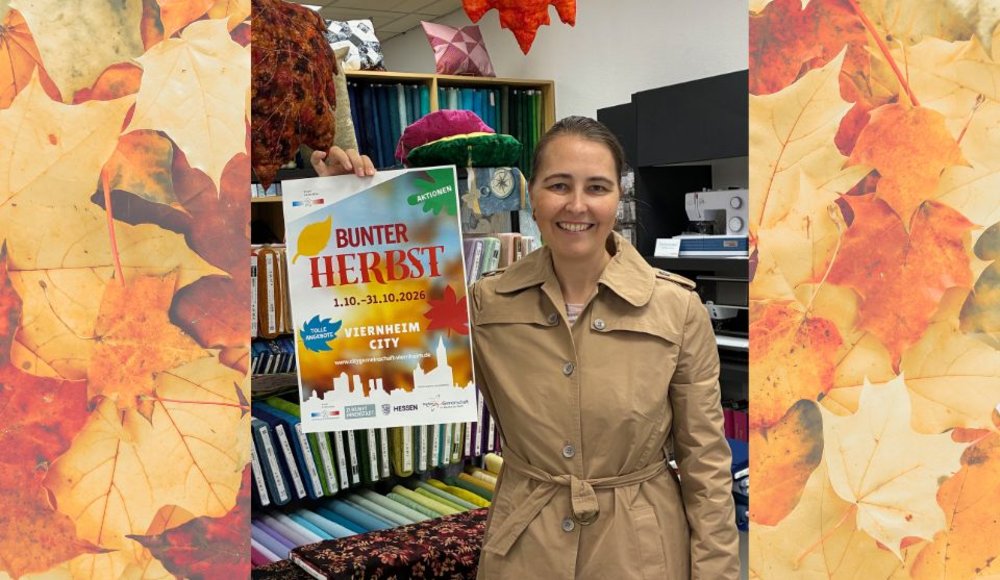 Renate Müller steht lächelnd in ihrer Nähgalerie und hält ein Plakat zur Aktion „Bunter Herbst“ in die Kamera. Im Geschäft sind herbstliche Stoffe und Dekorationen zu sehen.