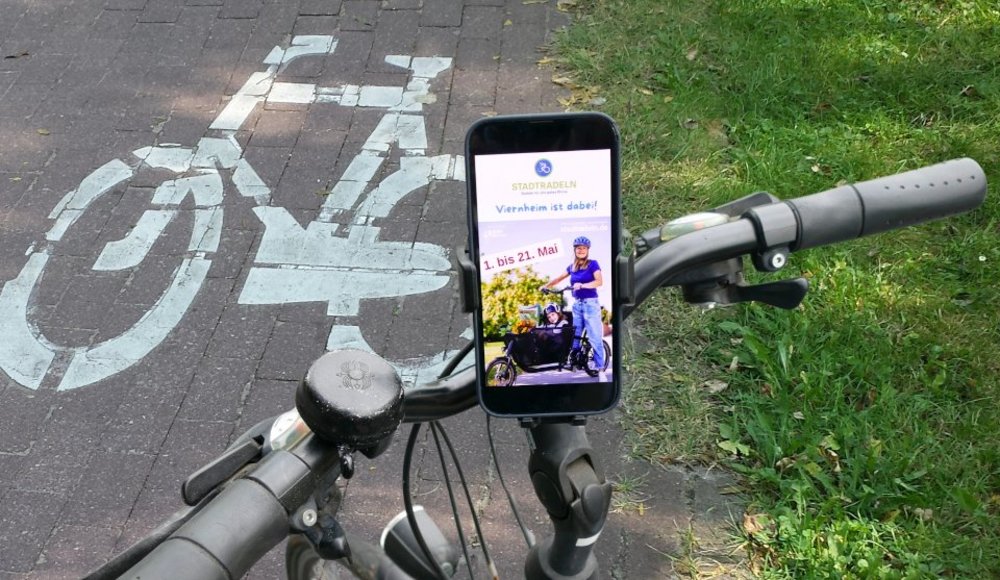 Fahrradlenker mit Handy in Halterung, darauf ist die APP zu sehen. Im Hintergrund die Straße mit Fahrradsymbol