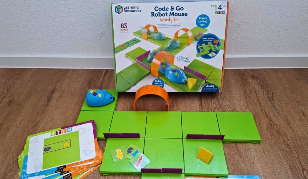 Aufgebautes Lernspiel mit einer blauen programmierbaren Robotermaus. Grüne Bodenplatten bilden einen Parcours mit kleinen Hindernissen und einem gelben Käse als Ziel. Daneben liegen Aufgabenkarten und Richtungskarten. Im Hintergrund steht die Verpackung des Lernspiels „Code & Go Robot Mouse“.