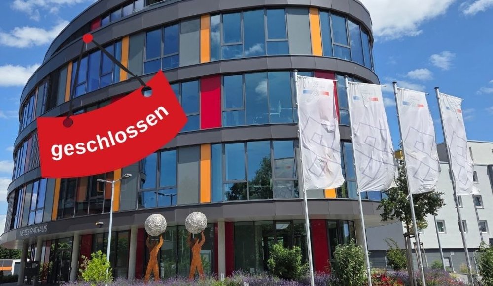 Zu sehen ist die Außenansicht des Neuen Rathauses in Viernheim mit seiner ovalen Form und der verglasten Fassade. Rechts wehen mehrere Fahnen mit dem Stadtlogo im Wind. Links weißt ein rotes Schild darauf hin, dass etwas geschlossen ist.
