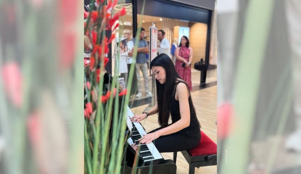 Eine junge Frau mit langen dunklen Haaren spielt Klavier im Rhein-Neckar-Zentrum. Im Hintergrund stehen mehrere Besucherinnen und Besucher, im Vordergrund sind hohe Pflanzen mit roten Blüten zu sehen.