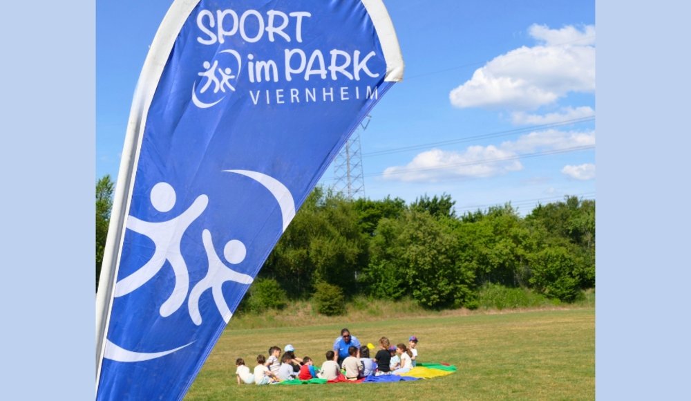 Blaues Banner mit der Aufschrift „Sport im Park Viernheim“ im Vordergrund; dahinter sitzen Kinder mit einer Betreuerin auf bunten Decken auf einer Wiese und nehmen an einem Sportangebot im Freien teil. Im Hintergrund sind Bäume und ein Strommast unter blauem Himmel zu sehen.